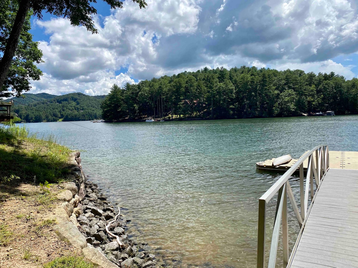 A serene view of Lake Glenville is presented, featuring calm water reflecting the sky. Lush, green trees frame the shoreline, while a private dock extends into the water. The surrounding hills contribute to the tranquil natural setting.