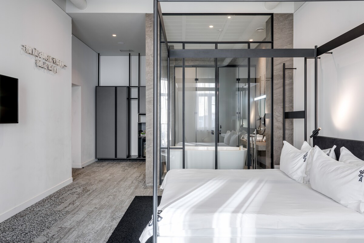 A modern bedroom features a spacious bed with crisp white linens, enclosed by a stylish metal frame. Large glass panels create a sense of openness, while a cozy seating area and a bathtub are visible in the background. Soft natural light fills the room.