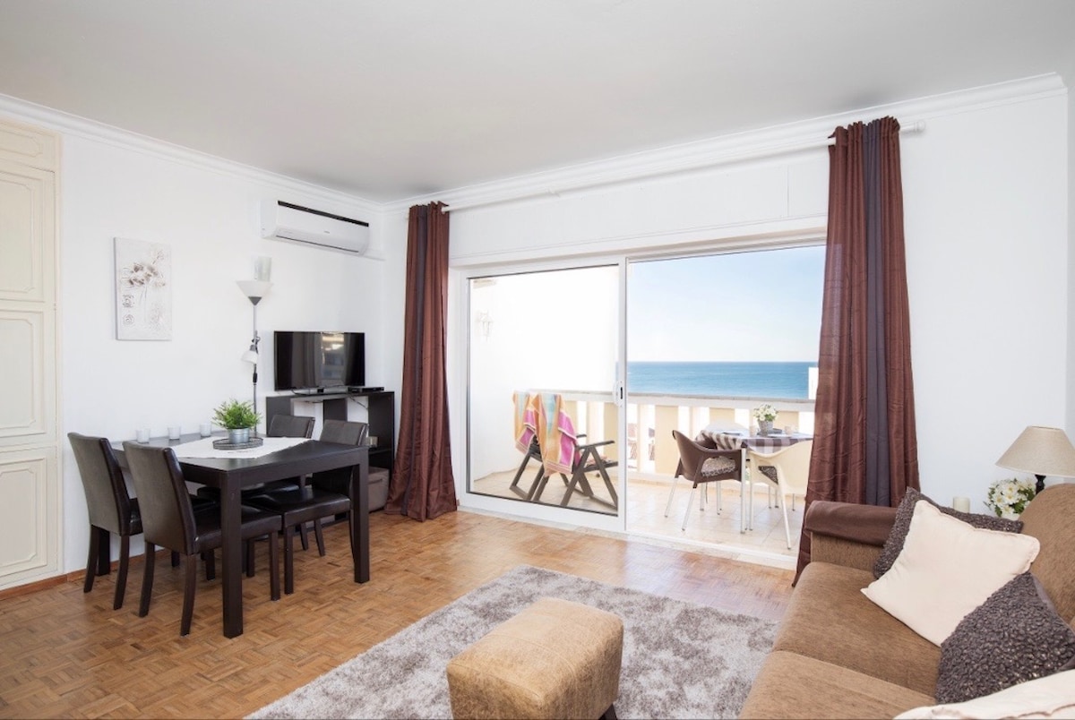 A spacious living area features a dining table with four chairs, a television, and a comfortable sofa. Large glass doors provide a view of the sea and lead to an outdoor terrace furnished with a table and chairs, perfect for enjoying the coastal scenery.