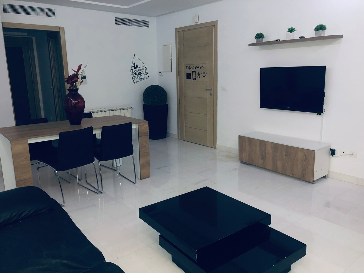 A modern living area features a rectangular dining table surrounded by four black chairs. A low black coffee table is positioned in front of a comfortable sofa. A wall-mounted television sits above a light wooden media unit, accented by decorative plants.