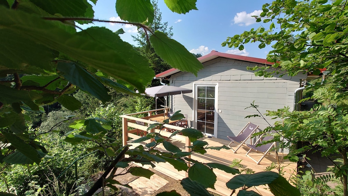 A cozy vacation home is surrounded by lush greenery, featuring a wooden deck with two lounge chairs and an umbrella. Large glass doors provide access to the interior, while plants frame the scene, creating a serene and private atmosphere.