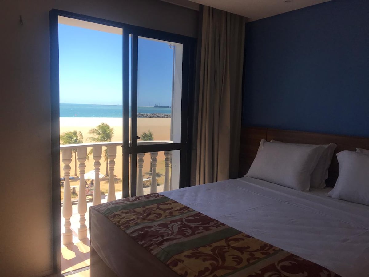 A comfortable bedroom features a spacious bed with crisp linens, illuminated by natural light from large windows. The view showcases the ocean and beach, emphasizing a serene coastal atmosphere. Light curtains frame the window, adding a touch of softness to the space.