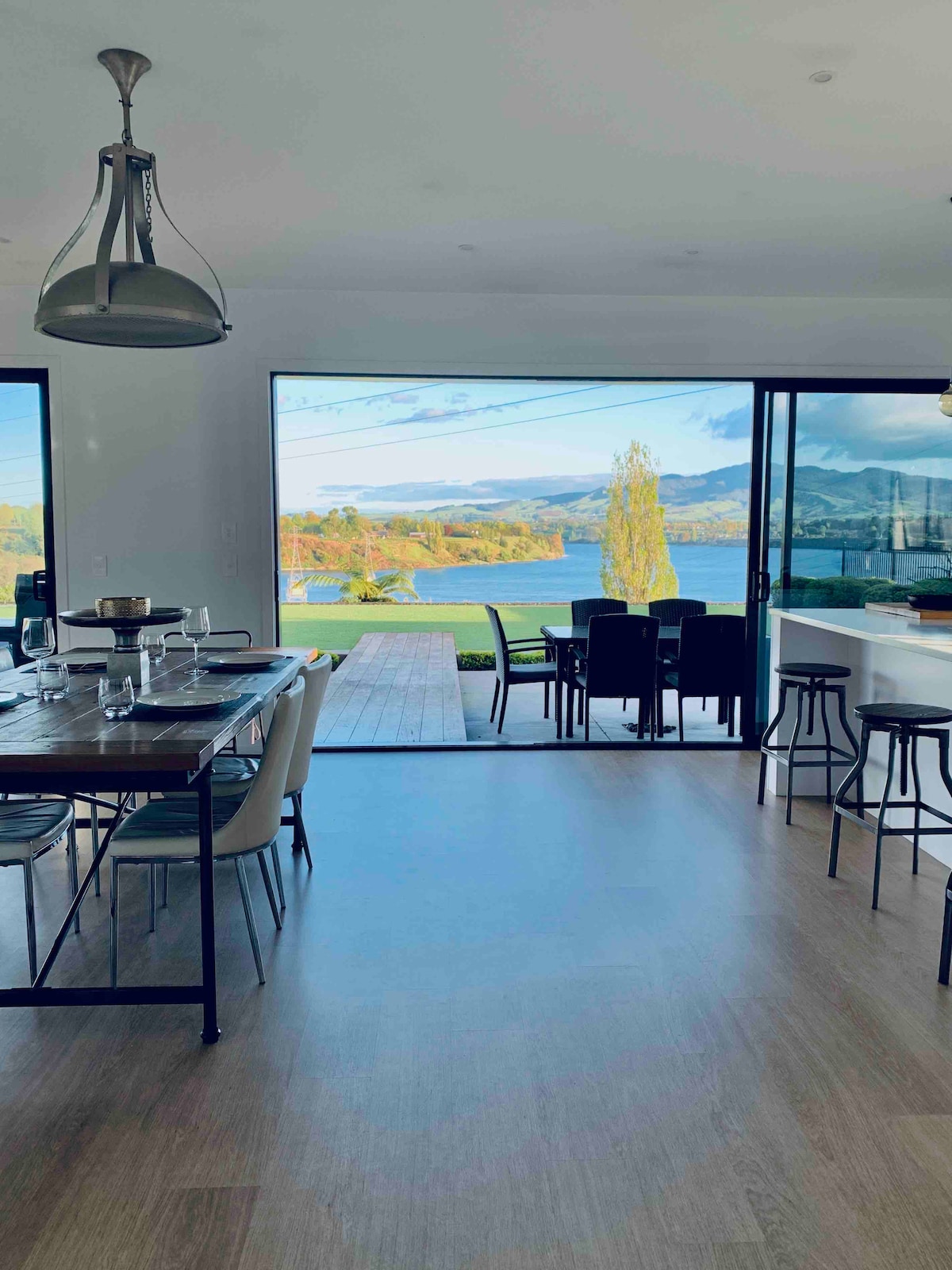 A modern dining area, featuring a wooden table set for meals, is framed by large glass doors that open to a scenic view of Lake Karapiro and mountains. Stools line the kitchen counter, and natural light brightens the spacious interior.