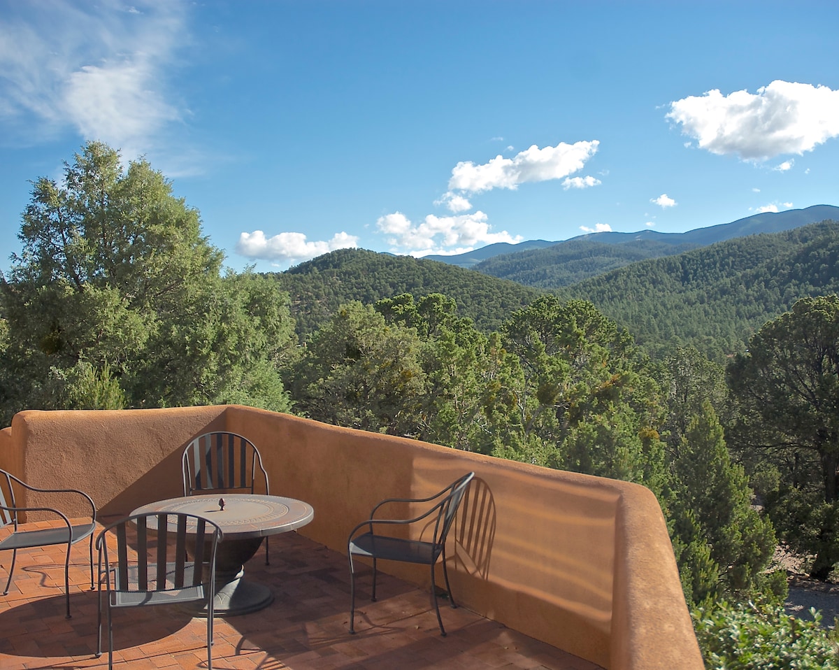 An outdoor patio area presents a circular table accompanied by four metal chairs. Surrounding trees enhance the natural setting, and expansive mountain views extend into the distance under a clear blue sky with scattered clouds.