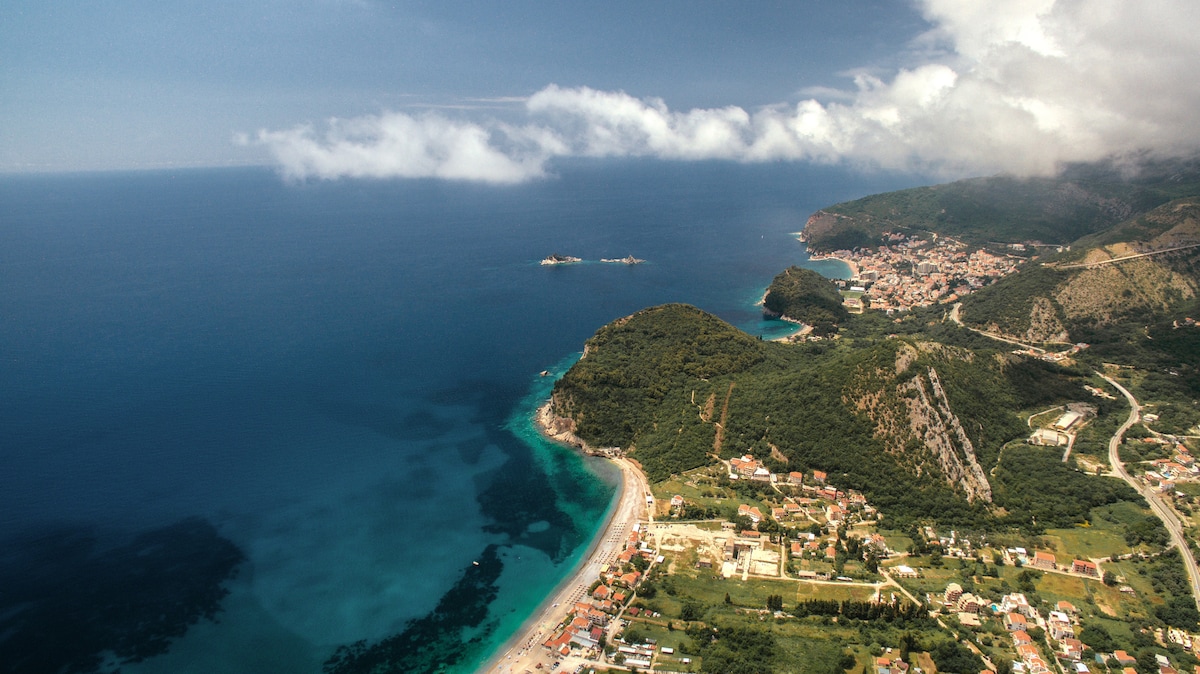 An aerial view captures the coastline of Buljarica, revealing a blend of green hills and turquoise waters. Beaches stretch along the shore, framed by rocky cliffs and scattered buildings. The distant horizon meets the sky, filled with scattered clouds, creating a serene coastal panorama.