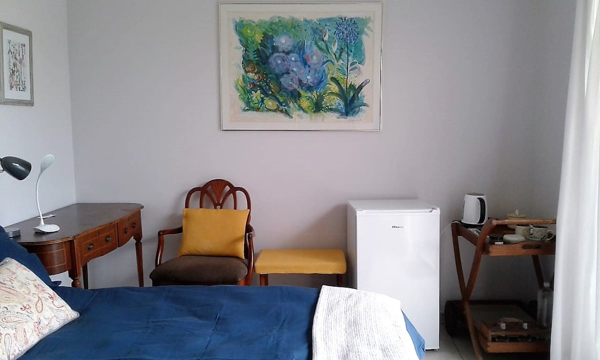 A cozy room features a spacious bed with navy bedding and decorative pillows. A wooden desk and chair are positioned beside a vibrant wall art piece. A bar fridge, kettle, and small microwave oven are present, along with a wooden serving tray displaying tea items.