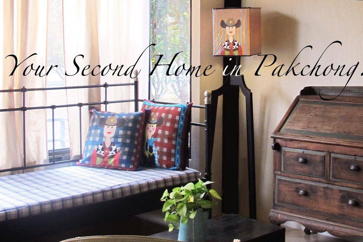 A cozy sitting area is highlighted by a wrought iron daybed topped with checked cushions. Natural light filters through sheer curtains, illuminating a wooden cabinet and a vibrant table lamp. A potted plant adds a touch of greenery to this inviting space.
