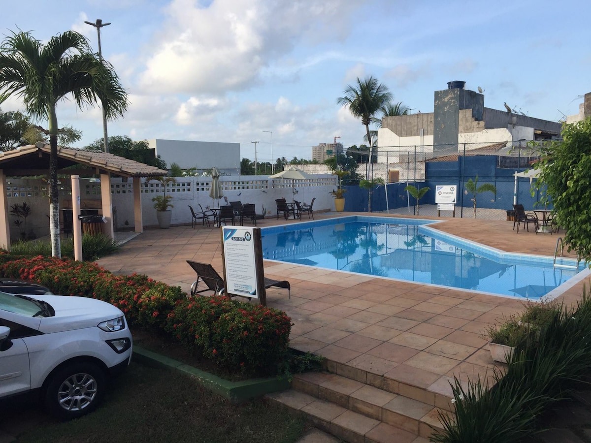 A well-maintained outdoor pool area is surrounded by lounge chairs and tropical foliage. A shaded seating area is visible near the pool, providing a relaxed space for gatherings. The pool reflects the sky, creating a tranquil atmosphere, while a sign indicates the pool rules.
