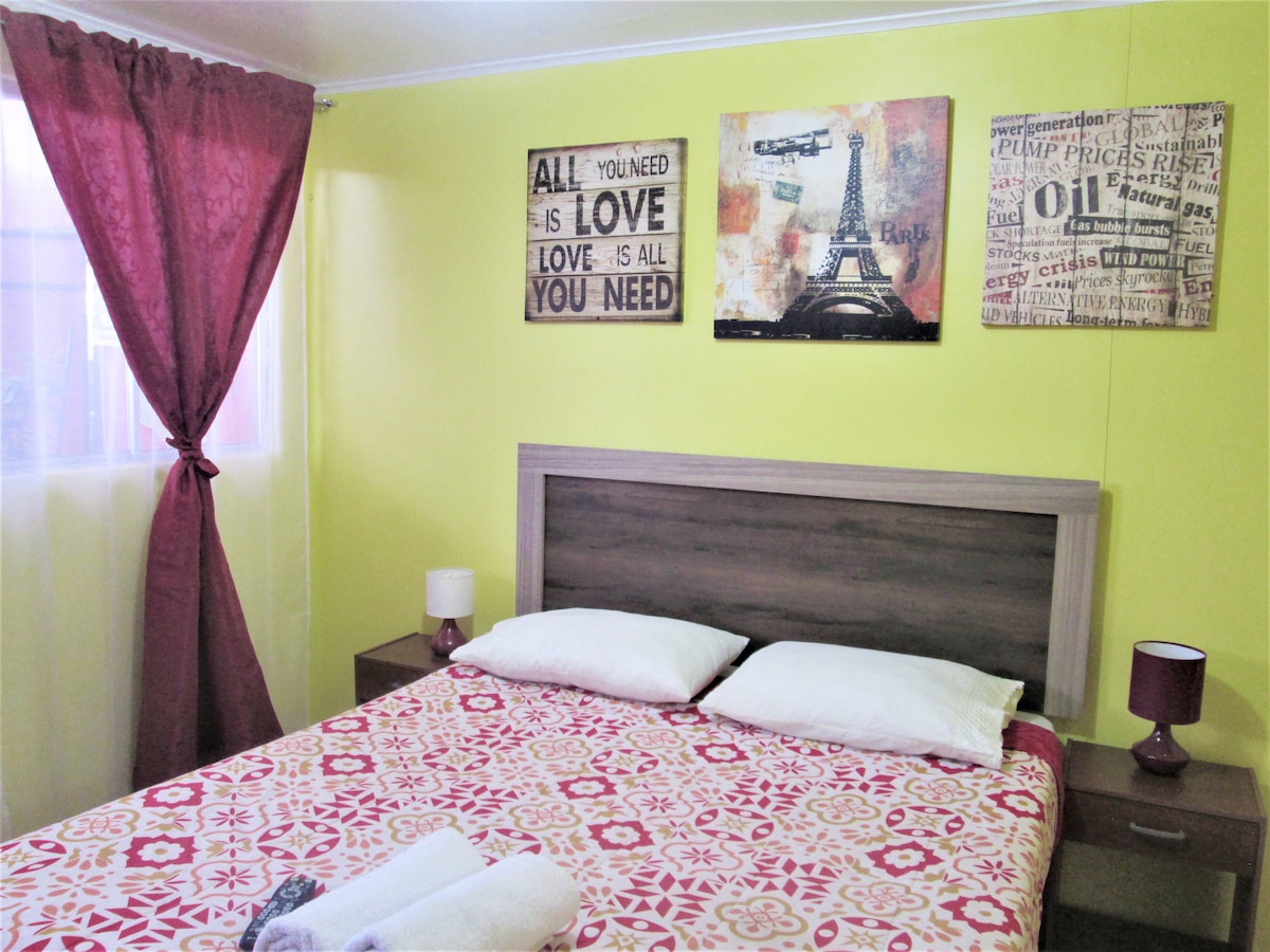 A well-lit sleeping area features a double bed with white linens and a patterned bedspread. Brown bedside tables with lamps are positioned on either side. Vibrant yellow walls are adorned with framed art pieces, and sunlight filters through a large window covered by sheer curtains.