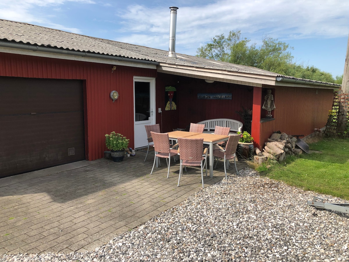 An outdoor seating area features a wooden table surrounded by several wicker chairs. The setting is complemented by potted plants and a garage door visible to one side. The red exterior of the building reflects a rustic charm against the backdrop of a clear sky.