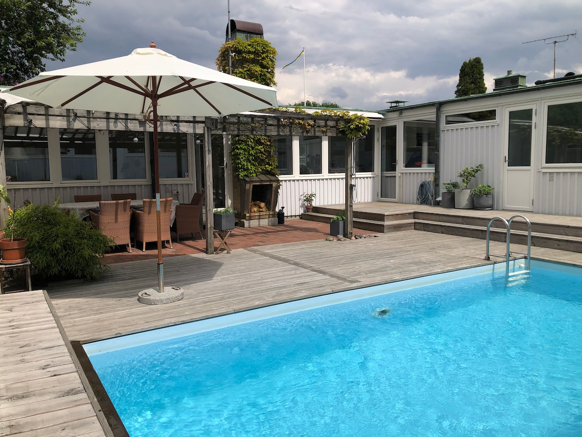 A fenced area is presented, featuring a clear blue swimming pool reflecting the sky. Comfortable seating is arranged near the pool under an umbrella. The surrounding deck is made of wood, complemented by greenery and planters along the edges, creating a serene outdoor space.