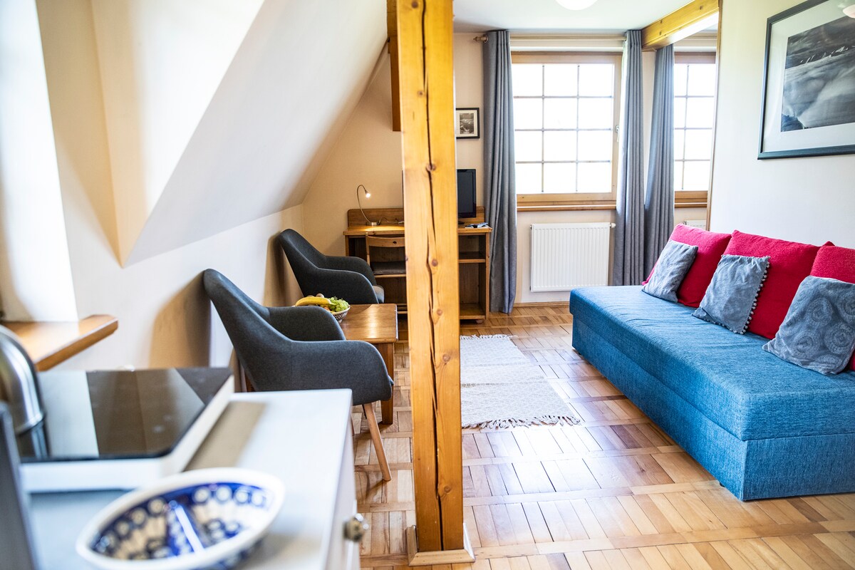 An airy living space features a spacious blue sofa adorned with red and gray pillows. Two comfortable chairs sit beside a wooden table, while natural light floods through the large window, framed by light gray curtains. Wooden beams add character to the ceiling.