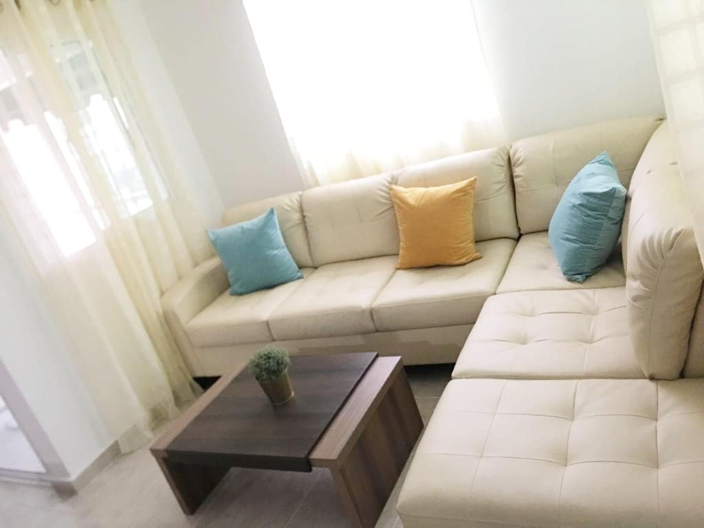 A light-filled living area features a cream-colored sectional sofa adorned with colorful throw pillows in blue and orange. A small wooden coffee table sits at the center, topped with a small green plant. Sheer curtains allow soft natural light to enter the space.