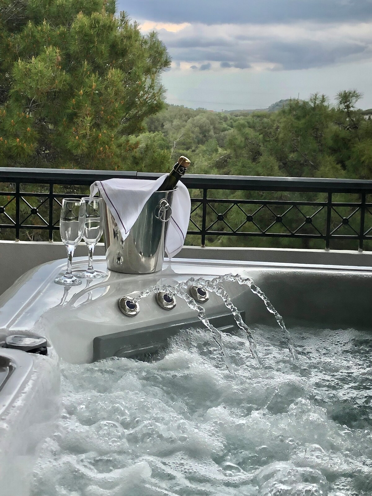 A hot tub is filled with bubbling water, accompanied by a silver bucket of champagne and two crystal glasses. The backdrop features a serene view of lush greenery, creating a peaceful environment inviting relaxation.