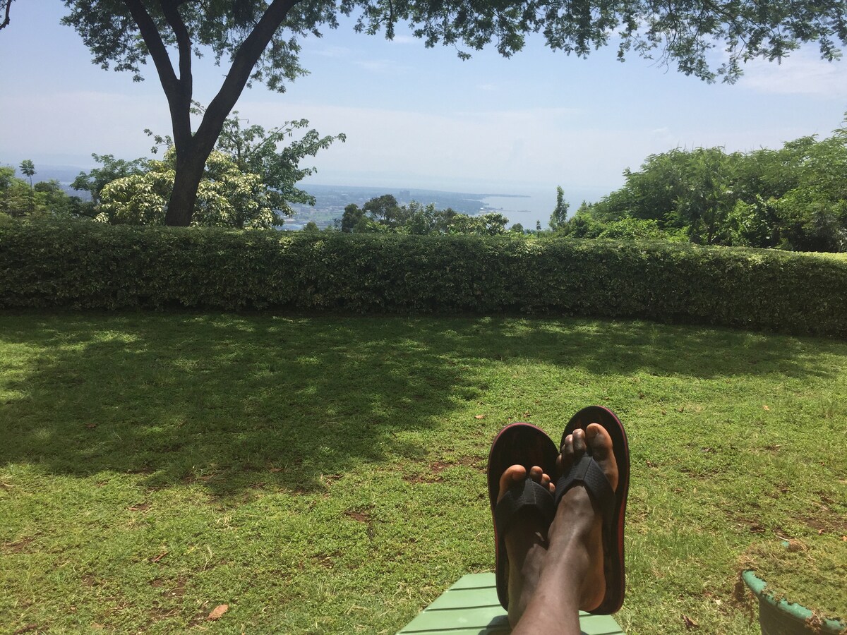 A serene outdoor space featuring a lush green lawn overlooking Lake Victoria. The scene includes a view of distant hills and trees, with a person relaxing on a lounger, their feet visible in comfortable sandals. The tranquil atmosphere is enhanced by the clear blue sky.