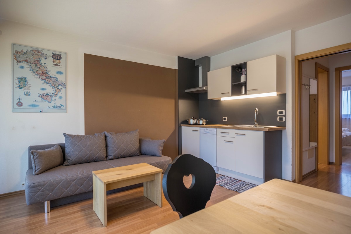 A cozy living space is featured, showcasing a sofa bed in a neutral grey tone, positioned alongside a minimalist coffee table. The kitchenette includes modern appliances and is highlighted by a contrasting dark wall. A wooden dining table with a uniquely designed chair completes the area.