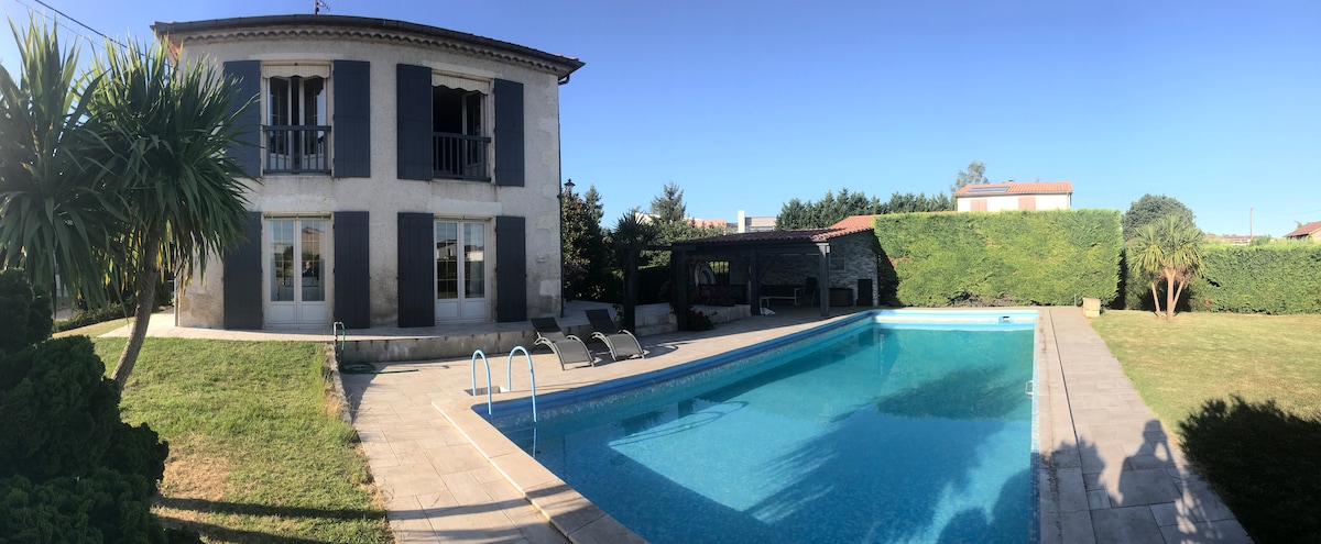 A panoramic view showcases a two-story house bordered by lush greenery. A swimming pool takes center stage, surrounded by a tiled patio and equipped with lounging chairs. The house features large windows and blue shutters, complementing the serene outdoor area.