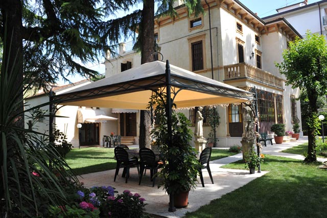 A charming garden area is featured with a gazebo providing shaded seating, surrounded by well-maintained greenery and colorful flower beds. The villa's elegant facade is visible in the background, enhancing the peaceful outdoor setting.