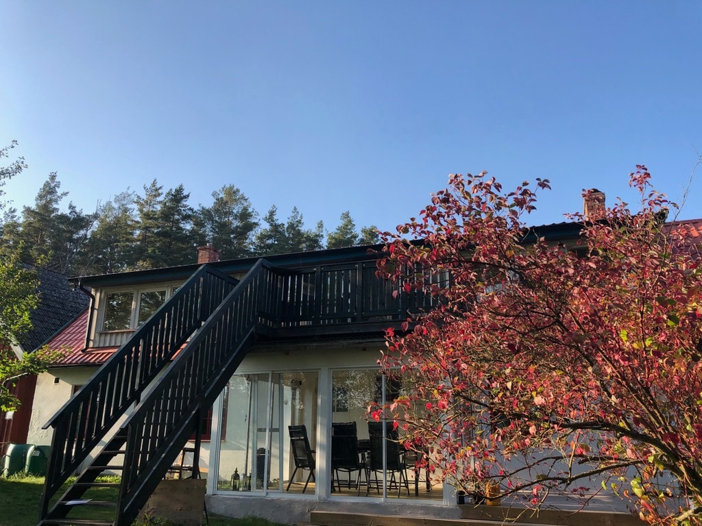 The exterior view captures a two-story structure with a wooden staircase leading to a balcony. A vibrant red tree adds a pop of color in the foreground, while the clear blue sky and forested area create a serene backdrop.