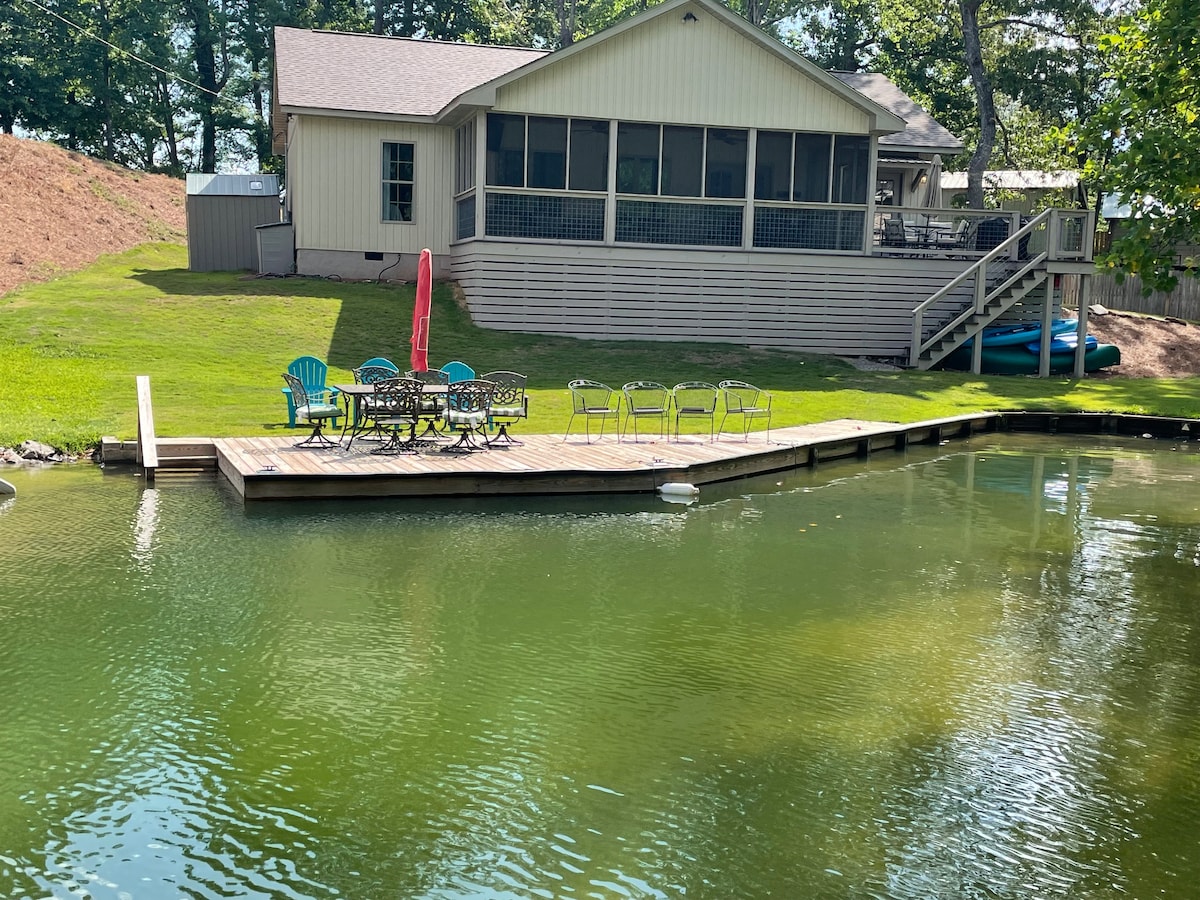 The exterior of a newly built cottage is displayed, featuring a large screened-in porch. A wooden dock extends into calm water, surrounded by several outdoor chairs and a table. Kayaks are visible nearby, with a grassy area leading to the cottage.