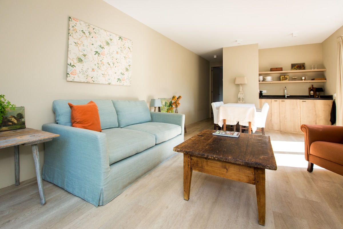 A light-filled living area features a soft blue sofa with an accent pillow and a rustic wooden table. A small dining space is set with a table and four chairs, while a modern kitchenette is visible in the background.