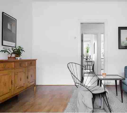 A bright living area features a wooden sideboard and a black wire chair beside a small table. A patterned rug covers the hardwood floor. A doorway leads to another room, hinting at the spaciousness of the layout.