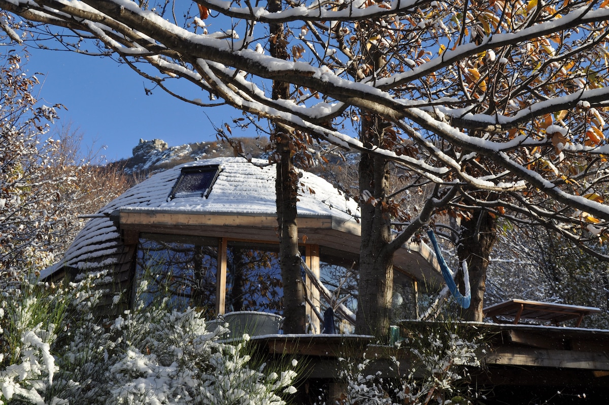 The spherical wooden house is enveloped in fresh snow, with a large skylight revealing part of the blue sky above. Surrounding trees are dusted with snow, and a wooden terrace extends from the home, providing a serene connection to the natural landscape.