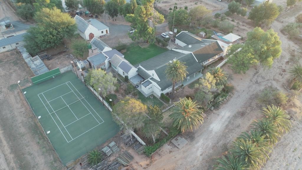 An aerial view of the property reveals a well-maintained homestead surrounded by greenery. A tennis court is visible in the foreground, alongside palm trees and various structures. The layout of the property suggests spacious outdoor areas, with plenty of open space for relaxation.