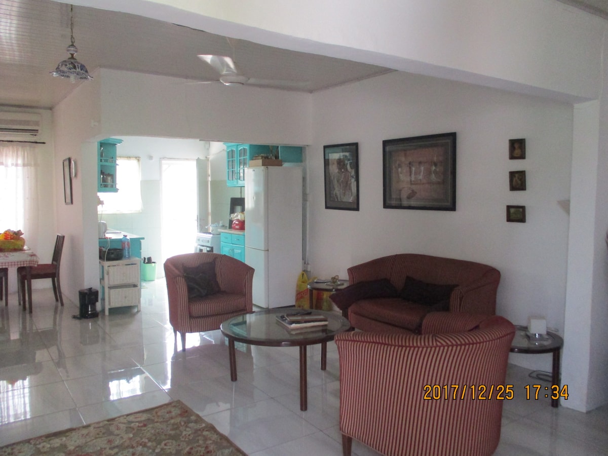 A spacious living area features two comfortable armchairs and a round glass coffee table. Light-colored walls enhance the bright atmosphere, with a vibrant rug underfoot. The open layout connects to a dining space and a kitchen, visible in the background, emphasizing a welcoming and functional environment.