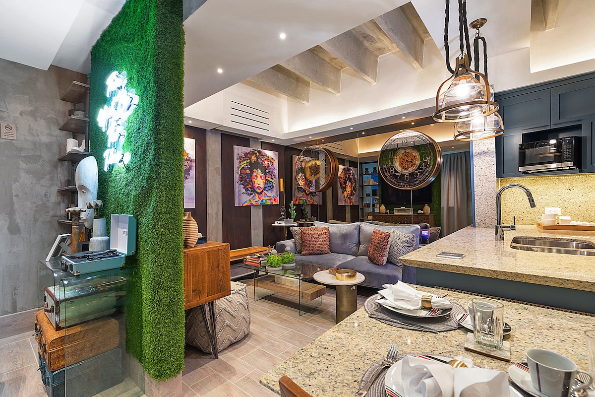 A contemporary living space features a mix of textures and materials. Green wall accents provide a natural touch, while a cozy seating area with colorful cushions invites relaxation. Artwork adorns the walls, and a well-equipped kitchen area offers modern appliances and stylish table settings.