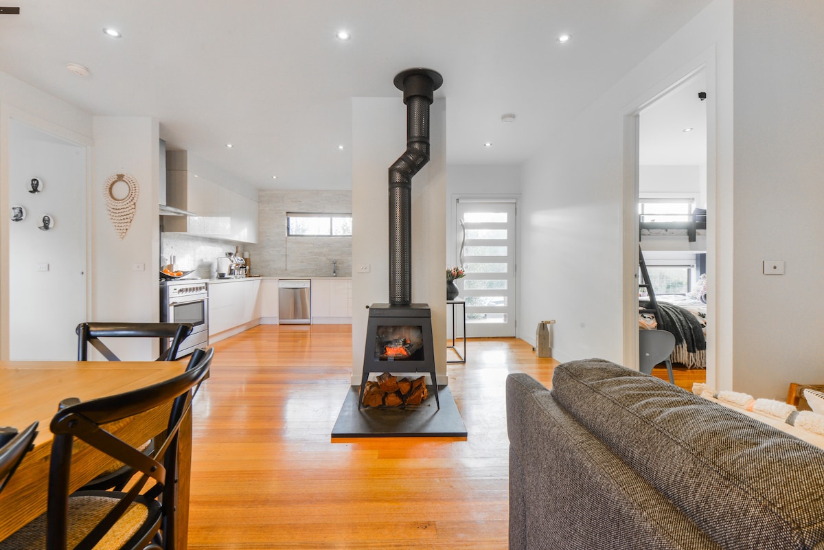 An open plan living area features wooden flooring and a cozy wood heater as a focal point. The kitchen, visible in the background, showcases modern appliances and cabinetry. Natural light floods the space through large windows, enhancing the inviting ambiance of the room.