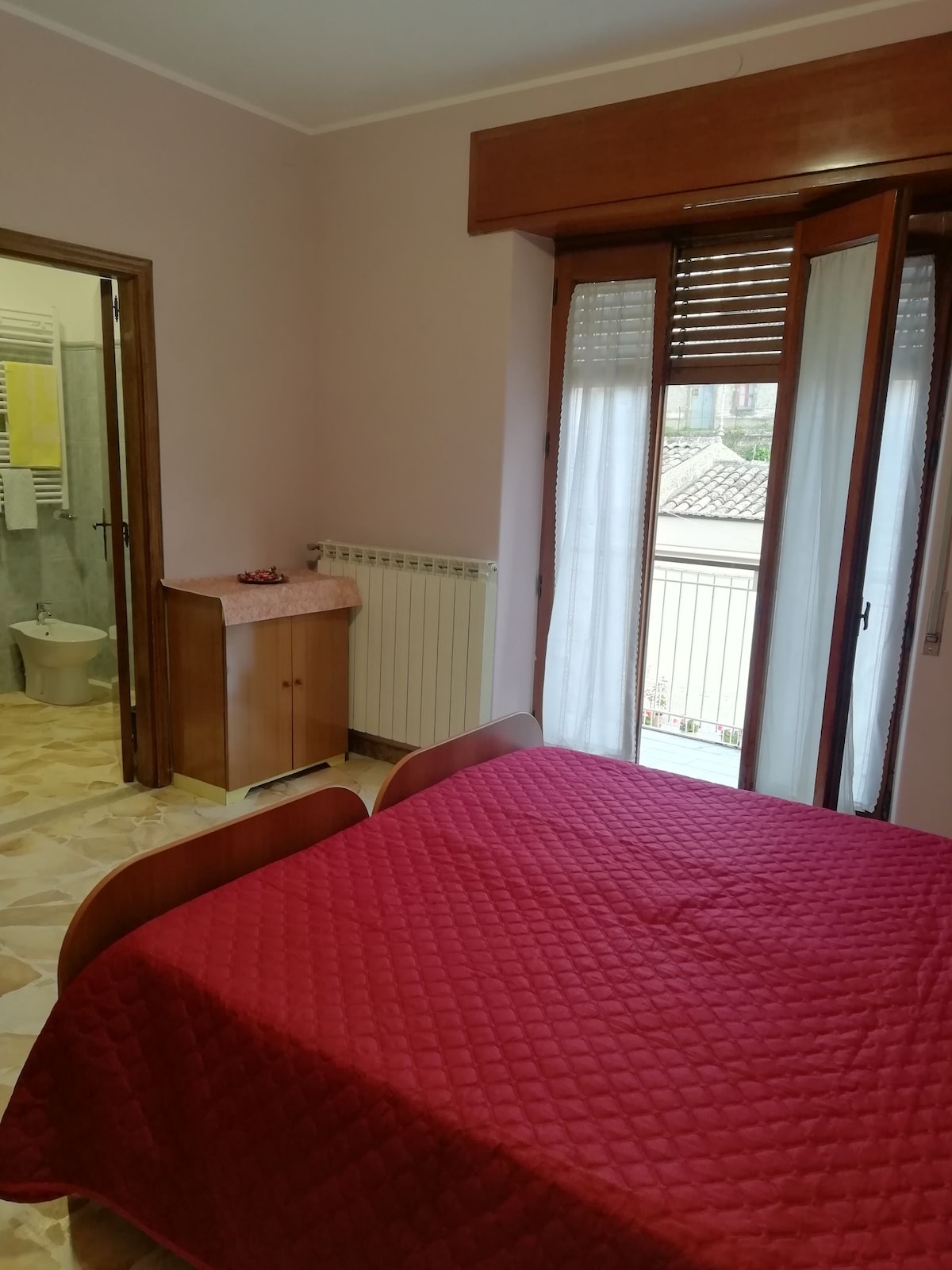 A softly lit bedroom features a spacious bed covered with a red quilt. Wooden furniture is present, including a cabinet. Light curtains frame the windows, providing glimpses of the outdoor view. An adjoining bathroom is visible through an open door.