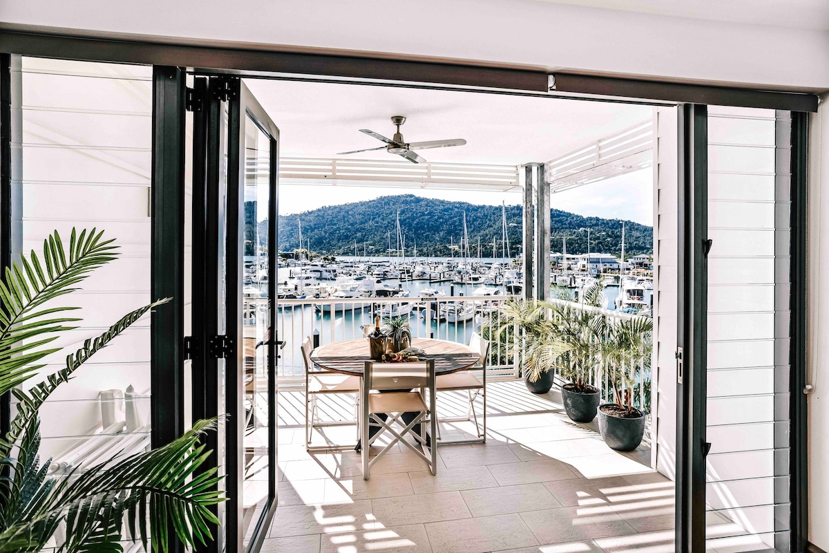 A spacious balcony features a large round dining table surrounded by potted greenery. Expansive sliding doors open up to reveal views of the marina and the distant hills beyond, with a ceiling fan providing comfort in the outdoor space.