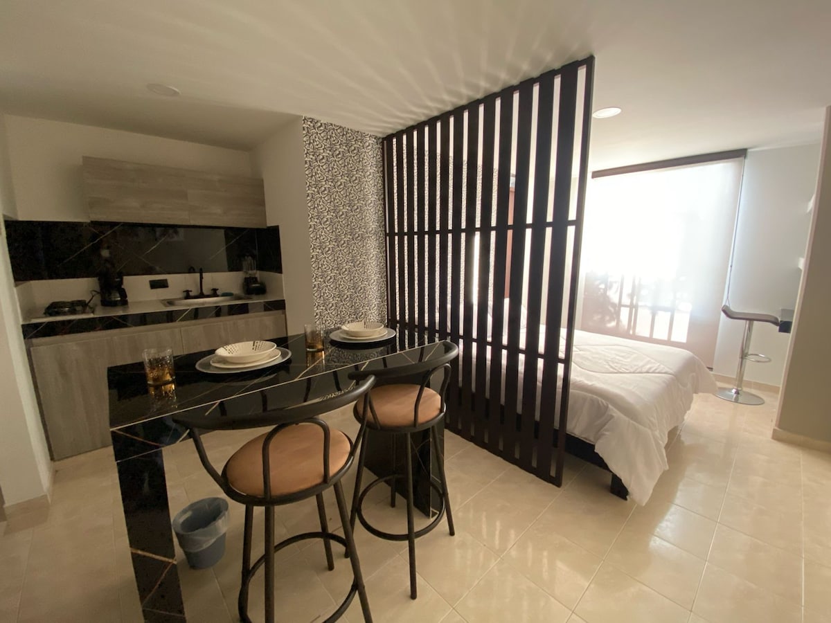 A modern space features a kitchen area with a black countertop and two high chairs, set next to a sleeping area separated by a patterned screen. Natural light fills the room, highlighting the white bedding on the double bed and the elegant design.