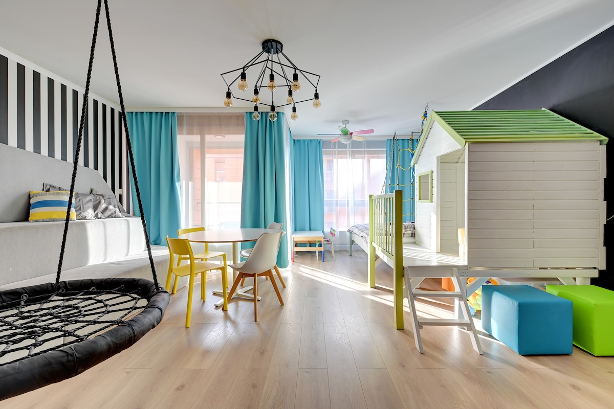 A bright living area features a large swing chair and a playhouse integrated with a bed. Vibrant blue curtains frame large windows, while a round table and colorful chairs enhance the space. Neutral wood flooring complements the playful atmosphere.