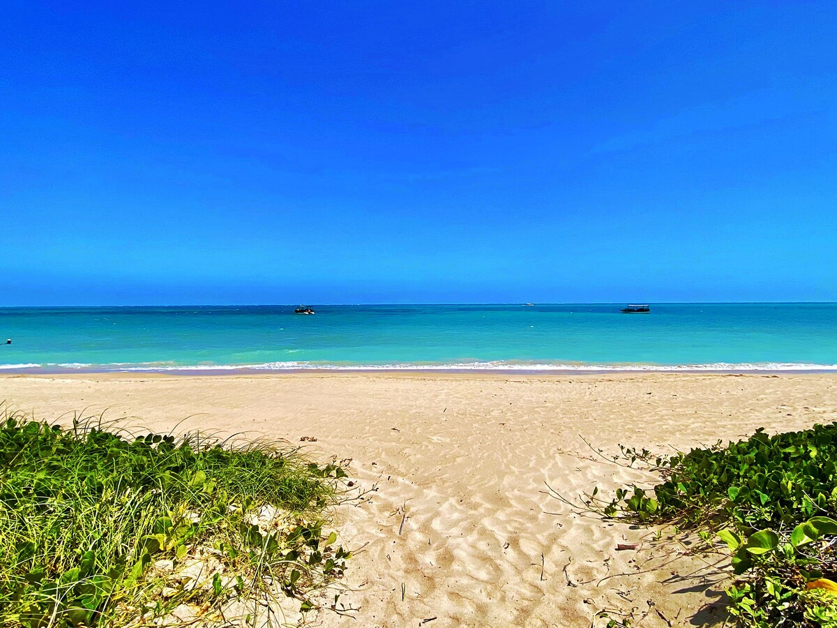 A sandy beach is presented, with a clear blue sky overhead and turquoise waters gently lapping at the shore. The scene is dotted with small boats on the horizon, while vibrant green vegetation edges the sand, enhancing the natural beauty of the coastline.