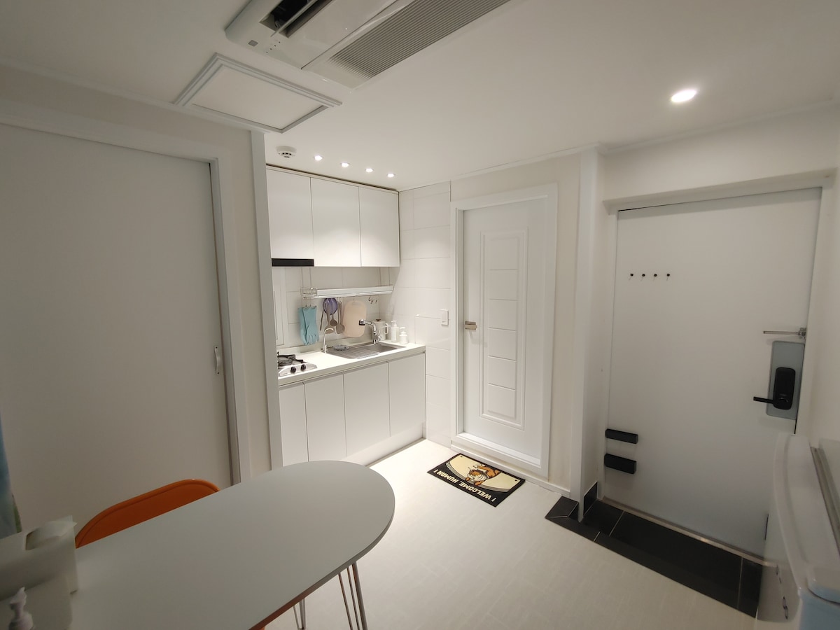 A modern kitchenette is featured with sleek cabinetry and counter space. Cooking essentials are neatly arranged, while a small dining table is placed close by. The entrance door with a secured lock is visible, along with white walls and bright lighting enhancing the space.