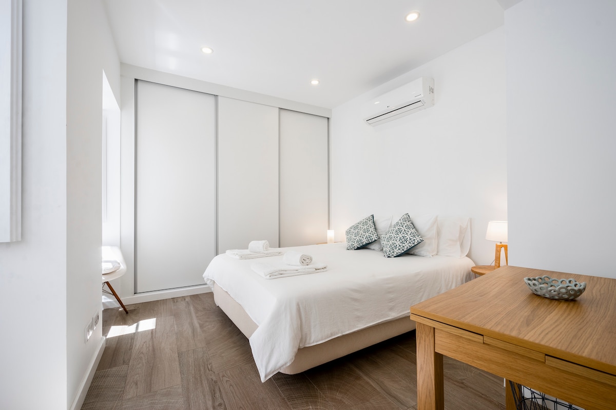 A bright and airy bedroom features a spacious bed centered against a white wall. Soft linens and decorative pillows are arranged neatly. A wooden table and bedside lamps are positioned beside the bed, while floor-to-ceiling sliding doors offer potential natural light.