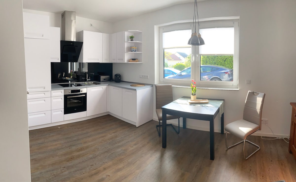 A modern kitchen is presented with sleek white cabinetry and a dark countertop. A dining table with two chairs is positioned near a large window, offering natural light and views of the outdoor area. Kitchen appliances and shelving add functionality to the space.