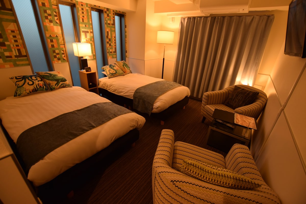 The room features two spacious beds topped with neutral-toned linens and decorative throws. Soft lighting creates a cozy ambiance. A striped armchair is positioned beside a small side table. Light filters through three windows, complemented by gray curtains.