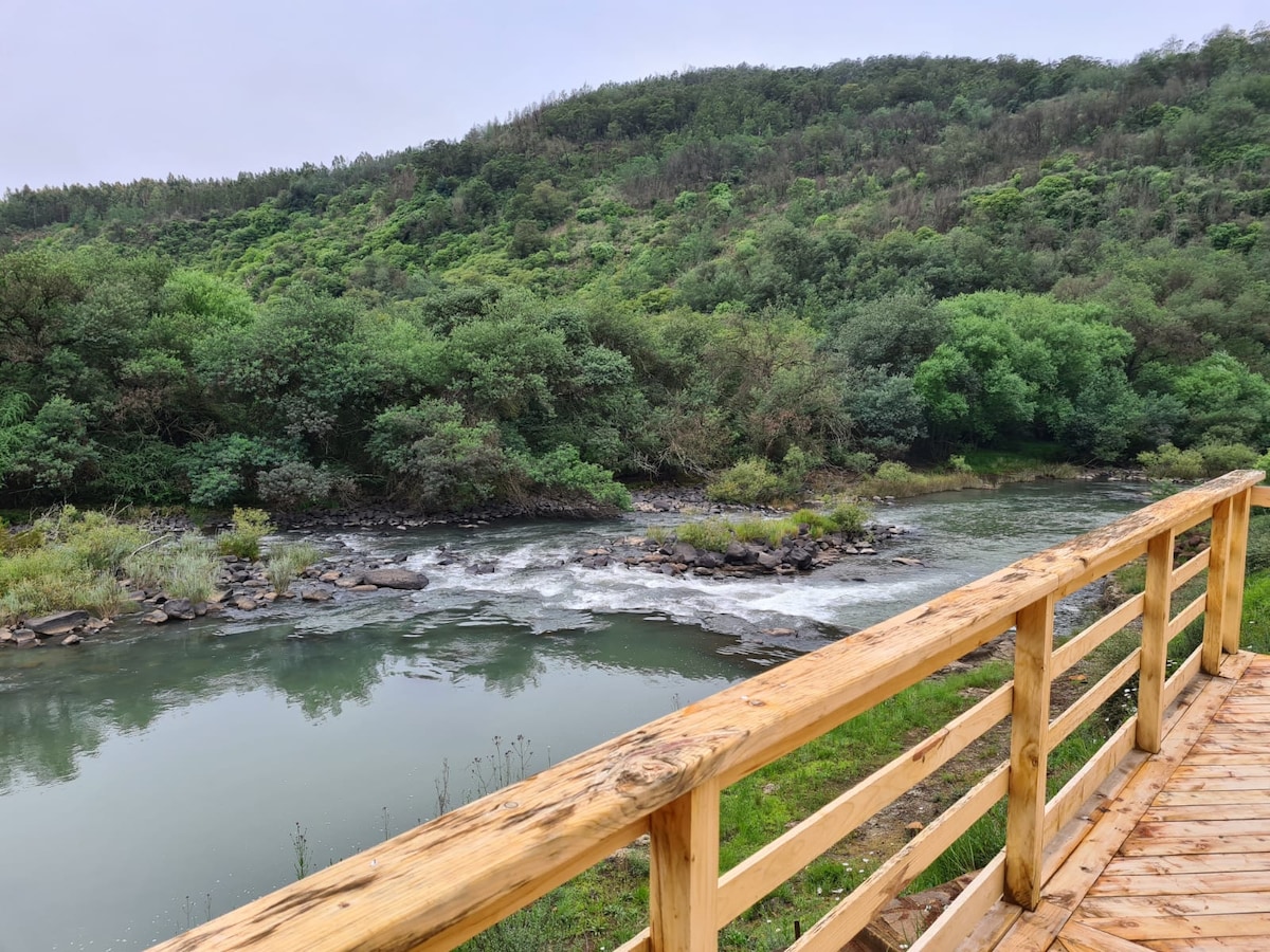 A wooden deck extends towards the Umzimkulu River, surrounded by lush green trees and rolling hills. The calm waters reflect the greenery, while gentle ripples can be seen where the river flows over rocks, creating a serene natural setting.