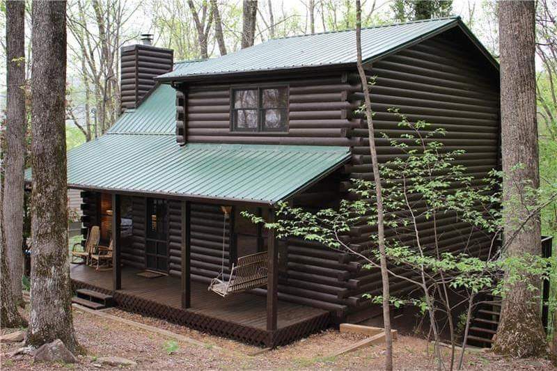 The rustic cabin, crafted with dark wood logs, features a green metal roof. A welcoming porch is equipped with a swing and two rocking chairs, nestled under the shade of surrounding trees. Large windows allow natural light to blend the interior with the outdoors.