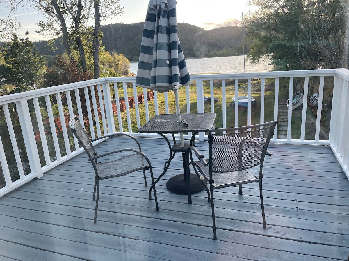 A small deck area is showcased, featuring a round table with a dark top and a single ashtray. Two metal chairs with mesh seating are positioned around the table. A striped umbrella provides shade, with a view of the lake visible in the background.