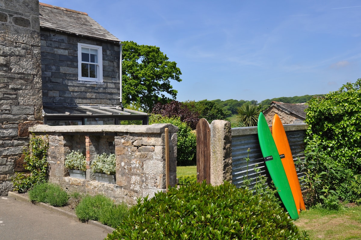 The exterior of Grove Cottage is depicted, featuring its traditional stone construction. Two surfboards in vibrant colors are propped against a stone wall, flanked by green foliage. A conservatory and lush garden can be seen in the background, contributing to the inviting atmosphere.
