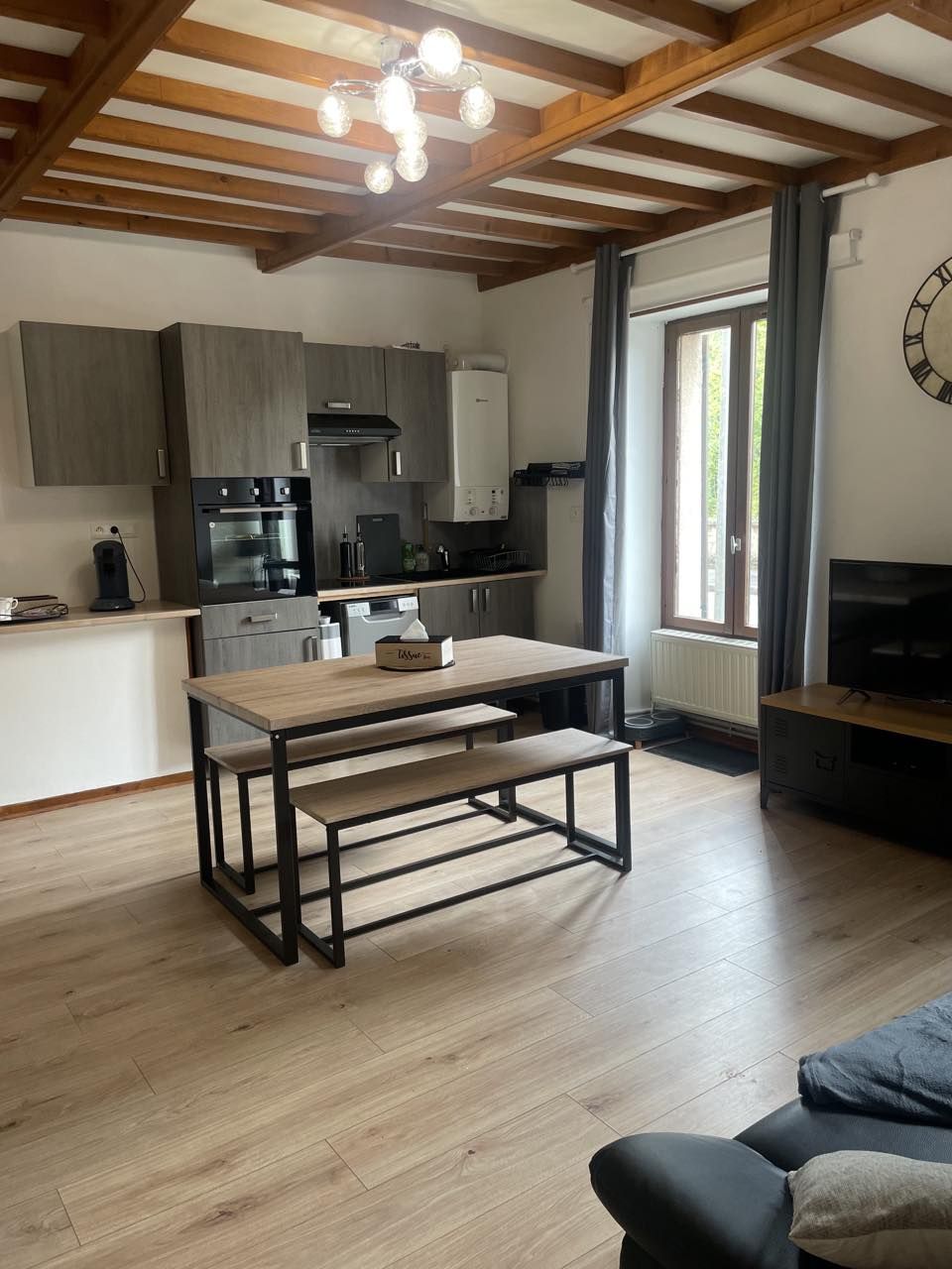 A spacious living area features a wooden dining table with benches, positioned near a window that allows natural light to enter. The modern kitchen is equipped with sleek cabinetry and appliances. A TV sits in the corner, completing the inviting atmosphere of the room.