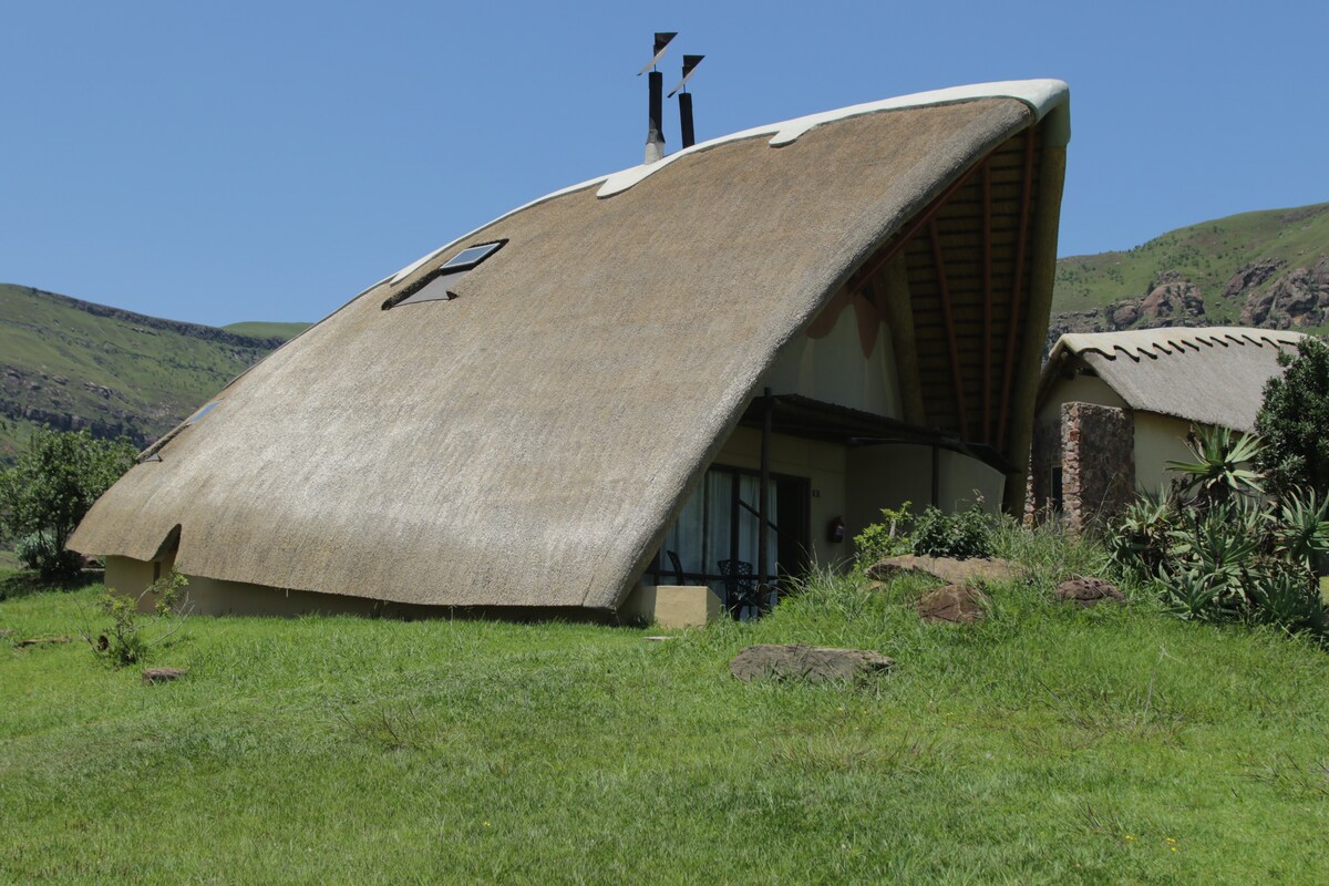 A unique cottage features a thatched roof with an angled design, blending harmoniously with the surrounding lush greenery. Large windows are incorporated to enhance natural light, while distant mountains provide a serene backdrop.
