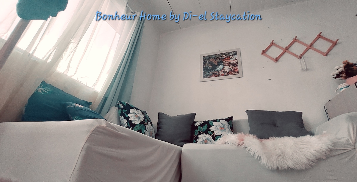 A cozy sitting area is featured, with light-colored sofas adorned with floral cushions. Soft textures are provided by a furry throw. Natural light filters in through large windows, enhancing the inviting atmosphere of the space.