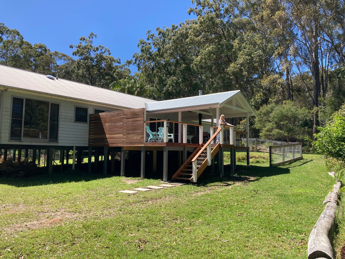 The cottage features an elevated deck with a wooden staircase leading down to the grassy yard. Light blue outdoor seating is visible on the deck, surrounded by lush greenery. The structure's exterior is painted in a light hue, complementing the natural surroundings.