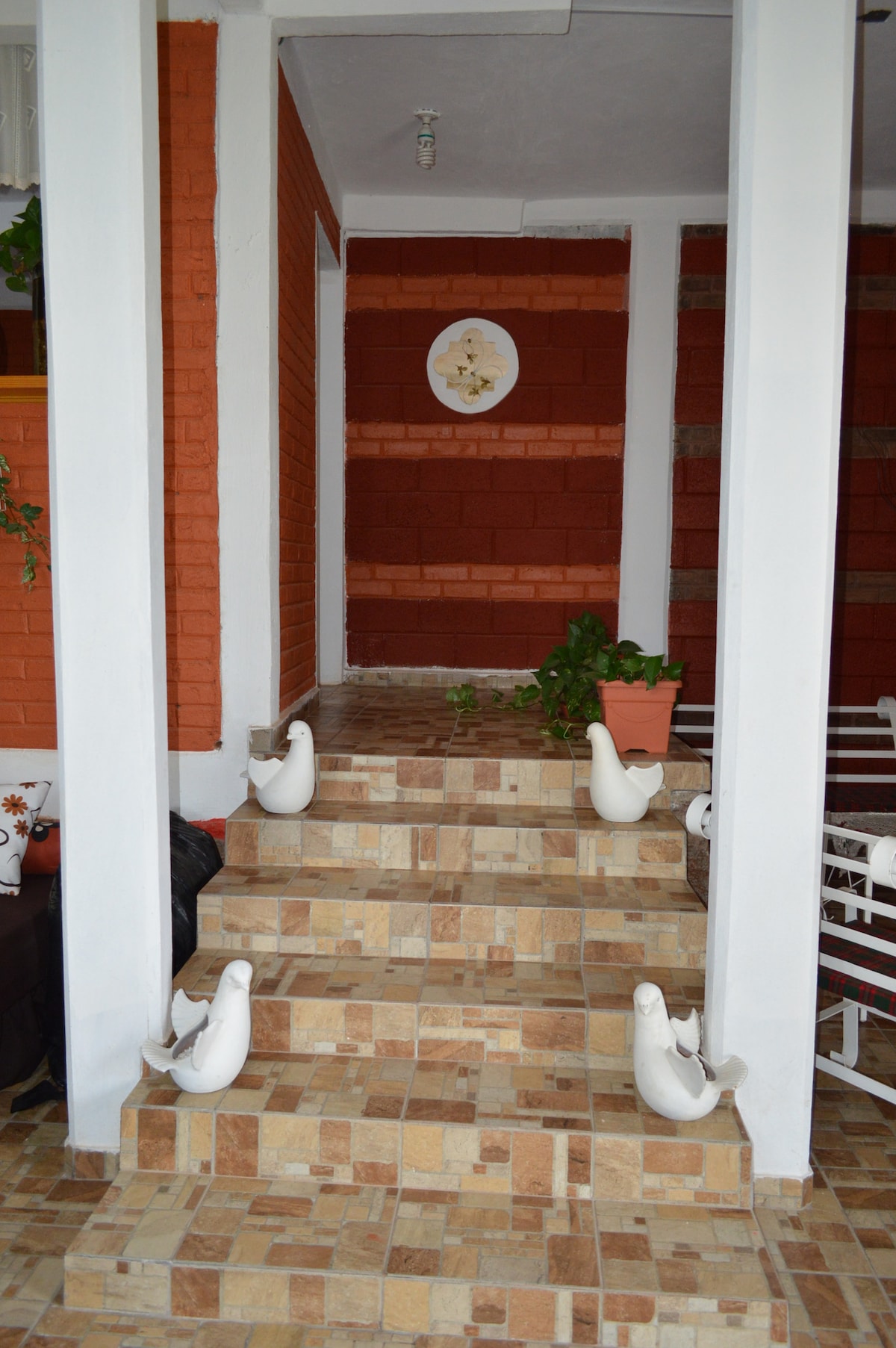 A set of stone steps leads upward, framed by white columns. The walls are painted in warm tones of orange and deep red. Decorative white sculptures are positioned on each side of the steps, with potted greenery adding a touch of nature to the entrance.