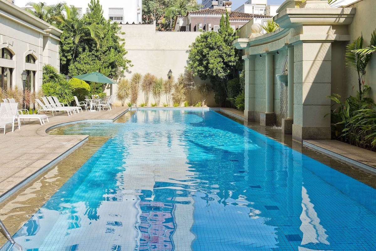 A large outdoor swimming pool is surrounded by landscaped greenery and a stone pathway. Lounge chairs are arranged along one side, while an umbrella provides shade at the far end. The water's surface reflects the surrounding structures and sky.